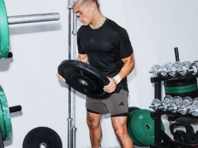 Ein junger Mann in einem schwarzen Hemd und grauen Shorts hebt eine schwere Langhantel in einem Fitnessstudio, das von verschiedenen Trainingsgeräten umgeben ist.