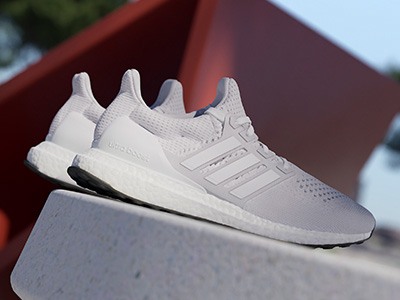 The image shows a pair of white Adidas Ultra Boost sneakers resting on a red background, with a blurred outdoor environment visible in the background.
