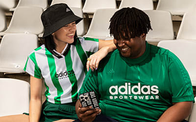 Two individuals wearing Adidas sportswear, one in a green shirt and the other in a black hat, are standing in a stadium-like setting with rows of white seats visible in the background.
