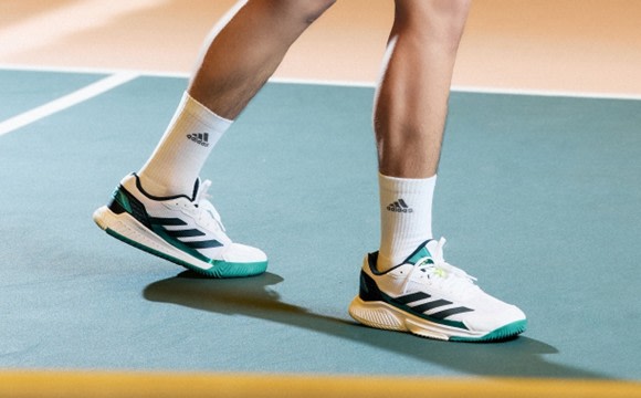 The image shows a person's lower body wearing white and green Adidas sneakers standing on a colorful sports court.