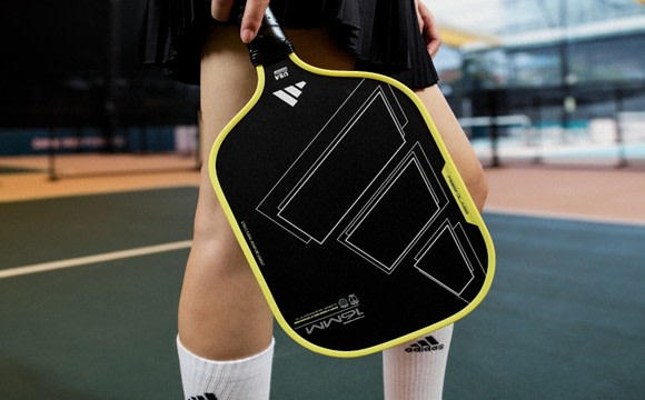 A person's hand holding a black and yellow tennis racket on a tennis court with a glass building in the background.