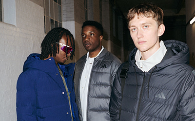 Trzy osoby, dwóch mężczyzn i jedna kobieta, stoją razem w otoczeniu, które wydaje się być miastem, z budynkiem widocznym w tle.