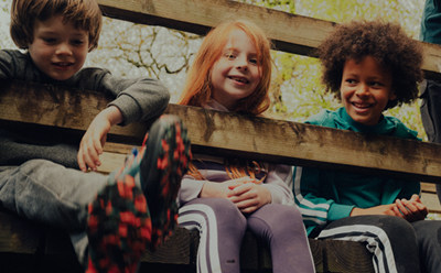 Tri deti, chlapec a dve dievčatá, sedia spolu na drevenej štruktúre vonku, obklopené stromami a listami v pozadí.