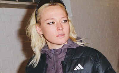 Mladá žena s blond vlasmi v čiernej bunde a fialovej košeli stojí na tom, čo sa zdá byť chodba alebo indoor prostredie.