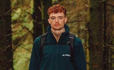 Młody mężczyzna z czerwonobrązowymi włosami i poważnym wyrazem twarzy stoi w leśnym otoczeniu, w zielonej kurtce i plecaku.