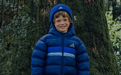 A young child wearing a blue hooded jacket is smiling while standing in front of a tree with lush greenery in the background.