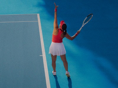 En person i en rød top og hvid nederdel spiller tennis på en tennisbane med en strålende blå himmel i baggrunden.
