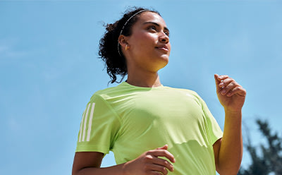 A young woman with curly hair wearing a bright yellow shirt stands against a clear blue sky, her expression thoughtful and serene.