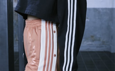 The image shows a person wearing a black jacket with white stripes and peach-colored pants with vertical stripes, standing in front of a blurred gray background.