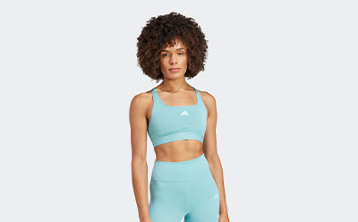 The image depicts a young woman with curly dark hair wearing a mint green sports bra and matching shorts, standing against a plain white background.