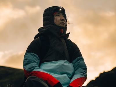 Una persona con una chaqueta negra con capucha y ropa verde y roja está de pie sobre un cielo nublado de fondo.