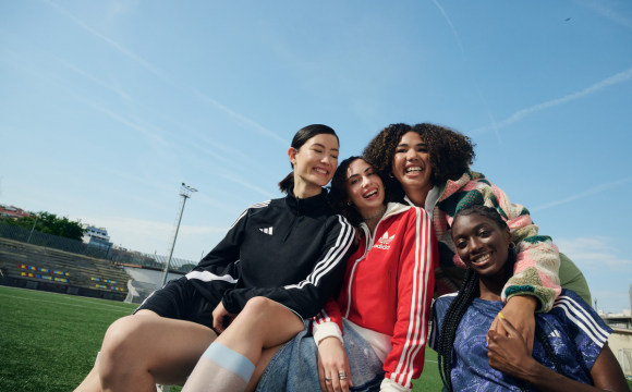 Two people wearing adidas running gear in front of a large, printed canvas depicting buildings in a city street. One is running, one is resting with their hands on their knees.