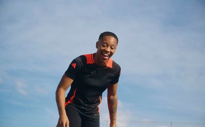 Um jovem usando uma camisa preta e suspensórios vermelhos está sorrindo e de pé contra um céu azul brilhante.