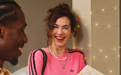 A young woman with curly hair and a bright smile, wearing a pink top, stands in front of a wall decorated with string lights.