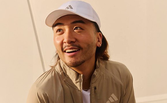 A smiling man wearing a white cap and beige jacket stands in front of a plain white wall.