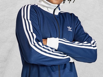 Das Bild zeigt eine Person, die einen dunkelblauen adidas Trainingsanzug mit weißen Streifen an den Ärmeln vor einem schlichten weißen Hintergrund trägt.