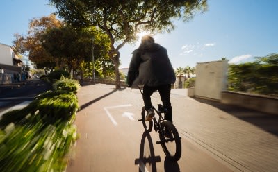 En person, der cykler på en asfalteret vej omgivet af træer og grønne områder, med en lys himmel i baggrunden.