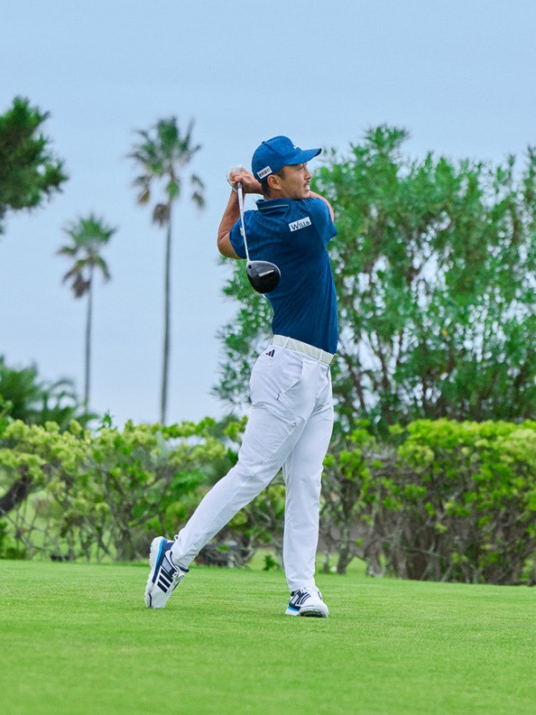 A person in a blue shirt and cap is swinging a golf club on a lush green golf course surrounded by palm trees.