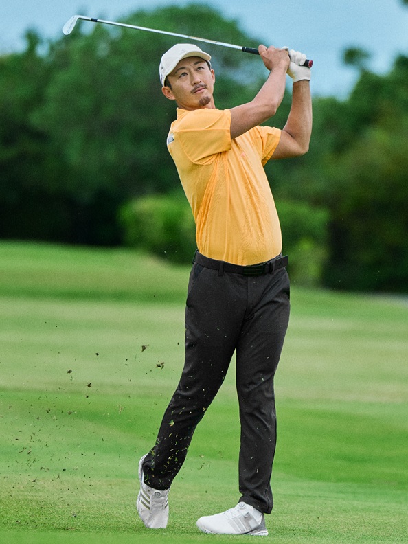 A golfer in a yellow shirt and black pants is swinging a golf club on a lush green golf course with trees and a clear sky in the background.