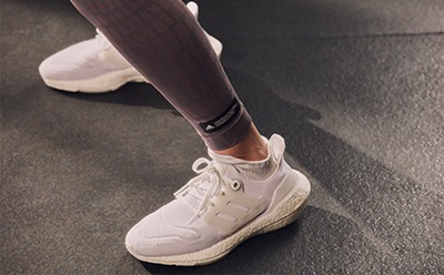 A pair of white sneakers with a person's legs wearing black leggings, standing on a gray floor.