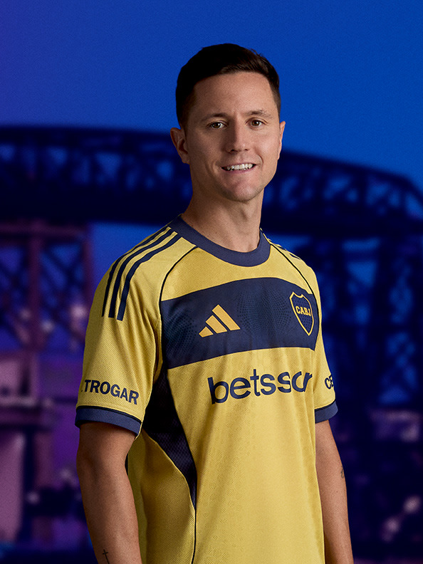 The image shows a smiling man wearing a yellow and black soccer jersey standing in front of a bridge at night.