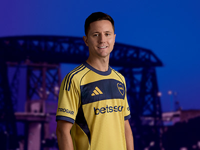A man wearing a yellow and black soccer jersey stands in front of a bridge at night, with a blue sky in the background.