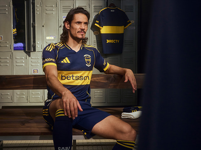 A man wearing a blue and yellow soccer jersey is sitting on a bench in a locker room, surrounded by lockers and other sports equipment.
