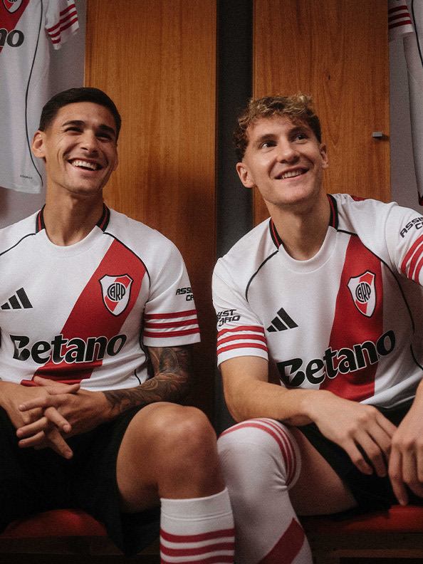 Two young men wearing white and red soccer jerseys with the "Betano" logo are sitting together and smiling in what appears to be a locker room or team facility.
