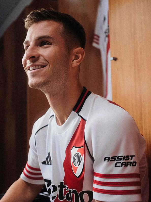 A smiling young man wearing a white sports jersey with red and black accents, standing in what appears to be a locker room or similar indoor setting.