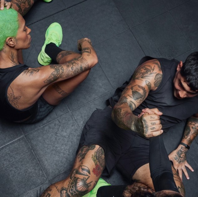 The image shows two individuals with extensive tattoos on their arms and upper bodies, engaged in what appears to be a physical altercation or training exercise on a dark, tiled floor.