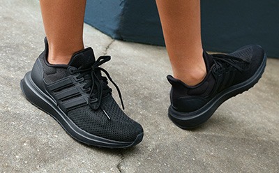 The image shows a pair of black athletic shoes with a mesh upper and a thick, textured sole, standing on a concrete surface.