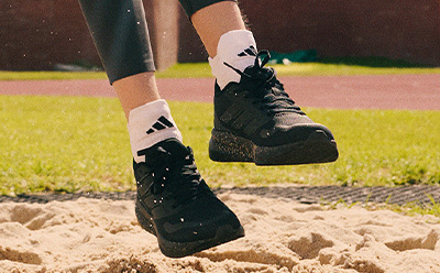 A pair of black and white athletic shoes are visible in the foreground, standing on a sandy surface with a grassy field in the background.