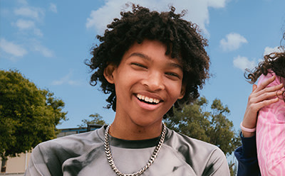 A smiling young person with curly hair stands in front of a background of trees and a blue sky.