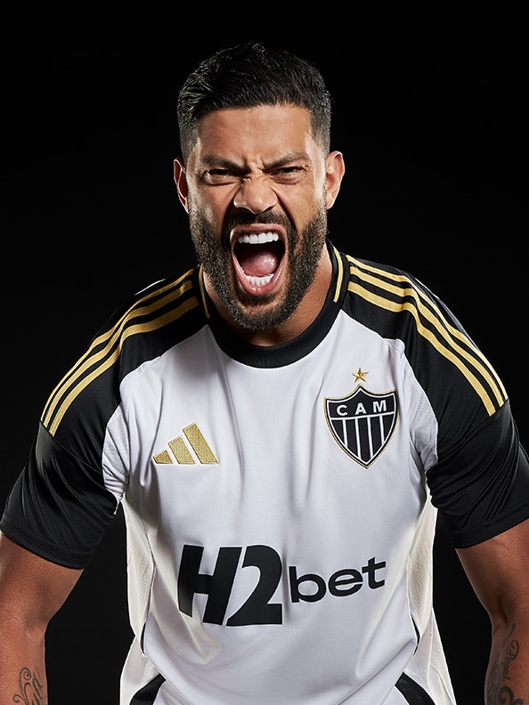A man with a beard wearing a white and black sports jersey is shouting with an intense expression on his face against a dark background.