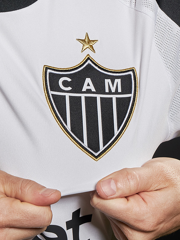 A white jersey with a black and gold emblem featuring a star and the letters "CAM" is being held by a person's hands.