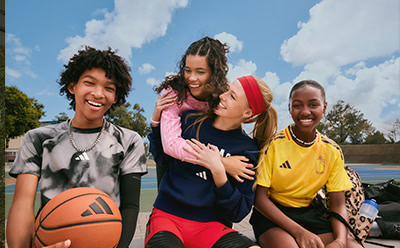 A group of young people, including two women and two men, are standing together and smiling on a basketball court with a blue sky and clouds in the background.