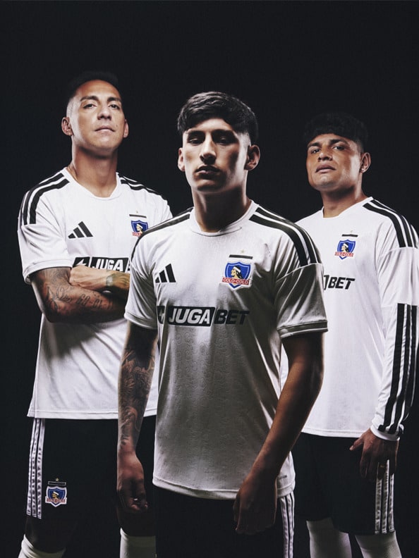 Three soccer players wearing white jerseys with the team logo and sponsor prominently displayed, standing against a dark background.