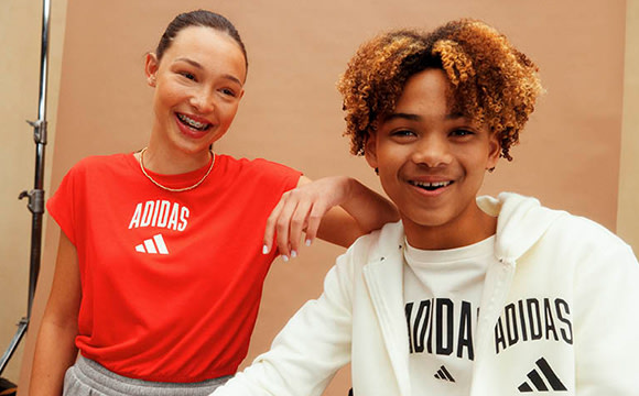 Two young people, a woman with long dark hair and a man with curly brown hair, are smiling and posing together in what appears to be a studio or indoor setting with a plain beige background.