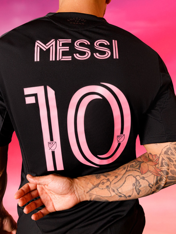 Na obrázku je osoba v černém dresu se jménem "MESSI"a číslem "10"natištěným na zádech na zářivě růžovém pozadí.