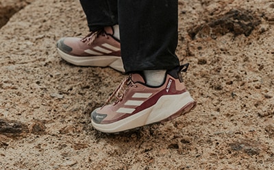 Das Bild zeigt die Füße einer Person, die ein Paar rosa-weiße Sneaker trägt und auf einer sandigen, felsigen Oberfläche steht.