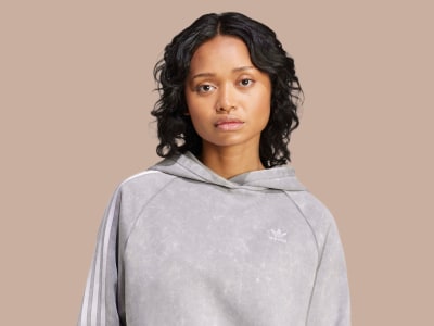 Una mujer joven de pelo oscuro rizado con una sudadera gris con capucha destaca sobre un fondo beige.