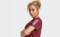 A young woman with curly blonde hair wearing a burgundy athletic top stands against a plain white background, her arms crossed and a confident expression on her face.