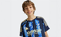A young boy with curly hair wearing an Adidas jersey is smiling at the camera against a plain white background.