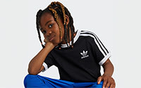 A young person with dreadlocks wearing a black Adidas shirt stands against a plain white background.