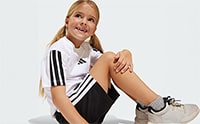A young girl with blonde pigtails wearing a white and black sports jersey is sitting on the floor and smiling at the camera.