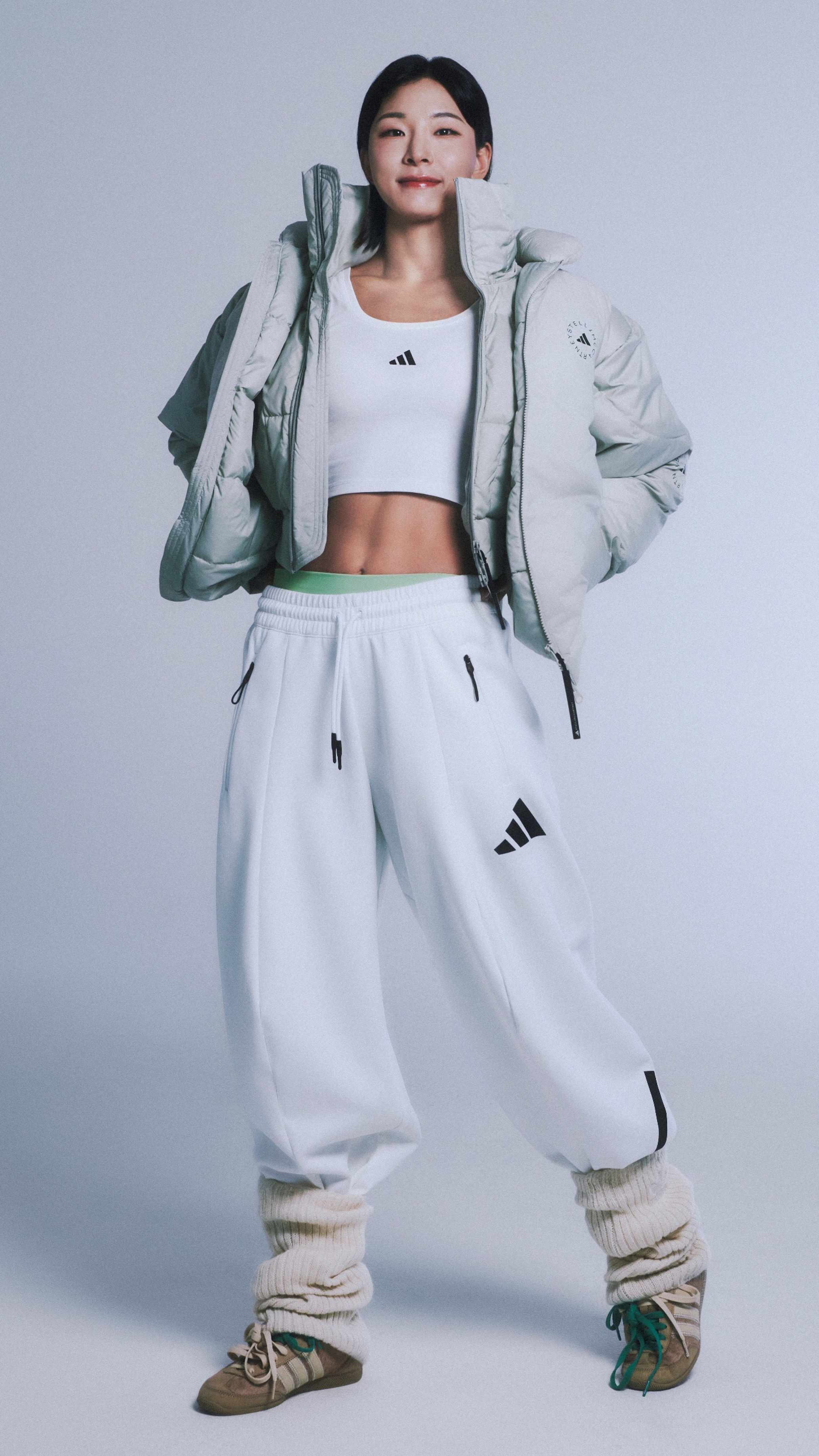 A young woman wearing a white crop top, a light-colored jacket, and loose white pants stands against a plain background, with her hair styled in a casual manner.