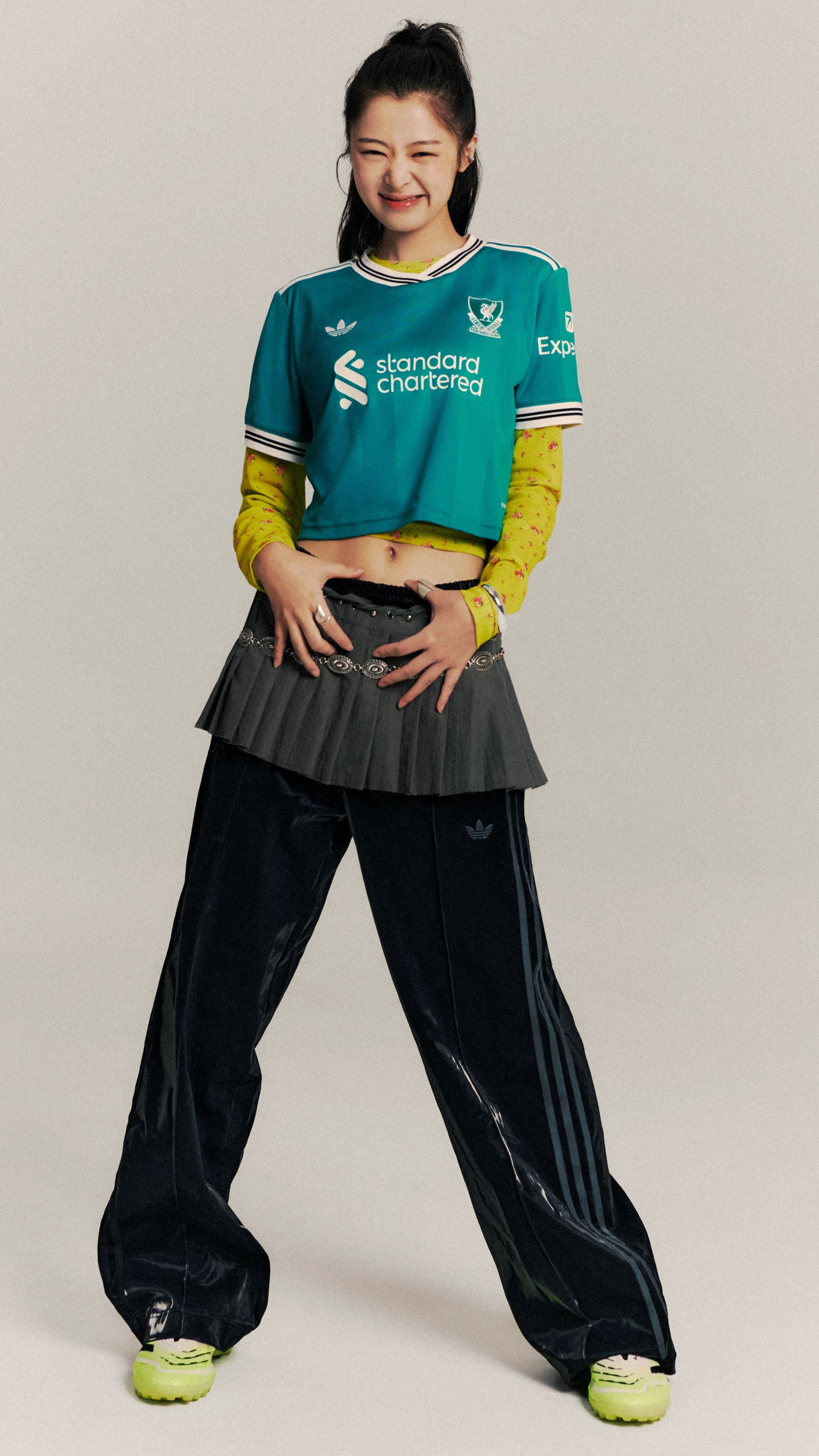 A young woman wearing a green and yellow sports jersey, black pants, and bright yellow shoes is standing with a confident and cheerful expression.