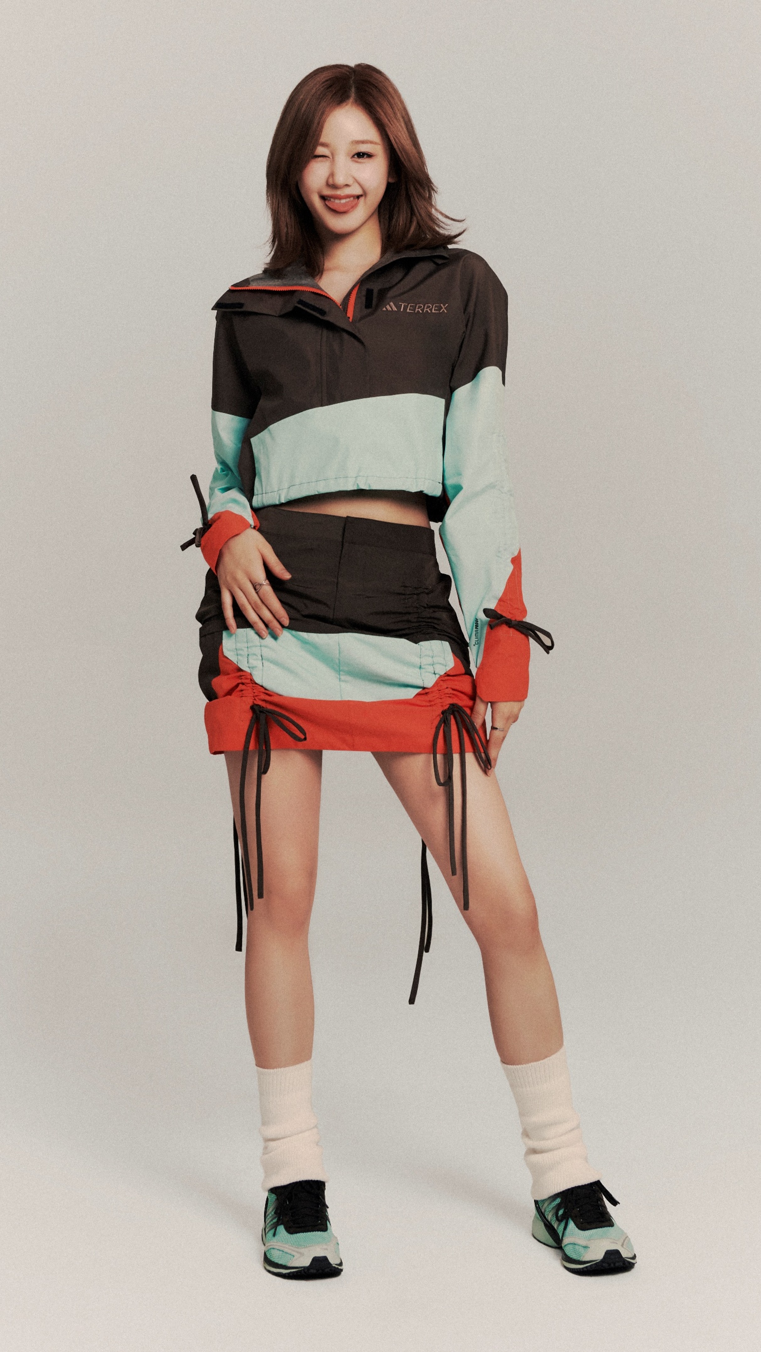 A young woman with long brown hair is smiling and wearing a colorful, layered outfit with a cropped jacket, shorts, and sneakers against a plain background.