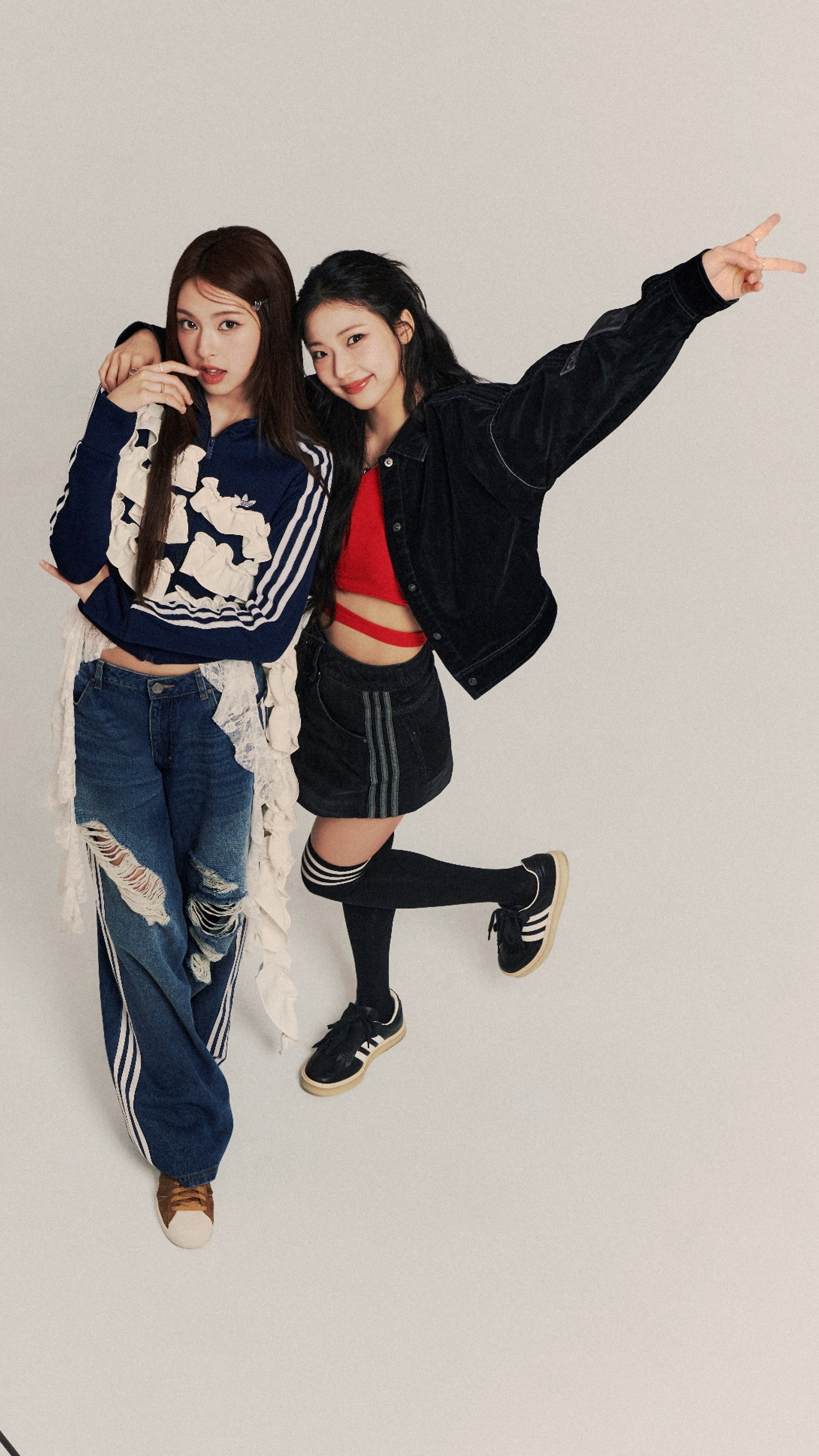 Two young women in casual clothing, one with her arm around the other, are posing and making a gesture with their hands against a plain background.