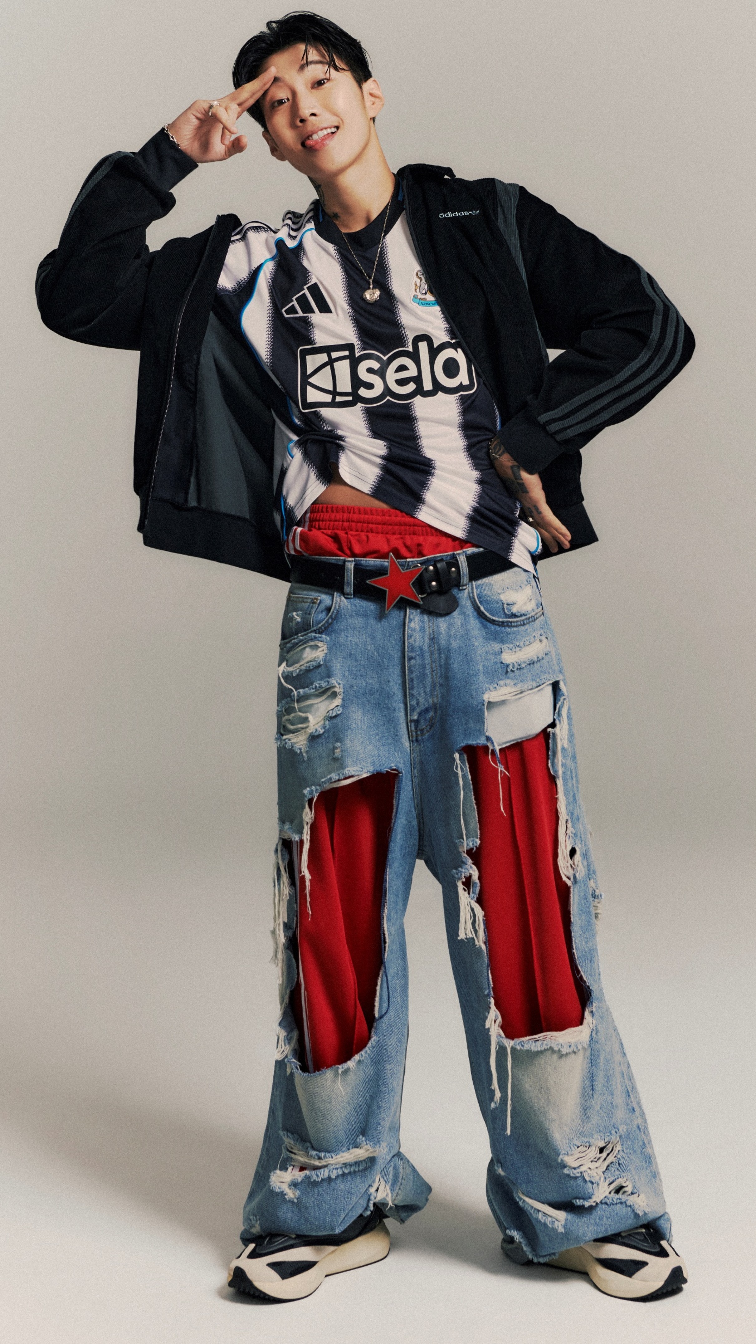 A person wearing a black jacket, a graphic t-shirt, and distressed jeans stands against a plain background, posing with their hands on their head.